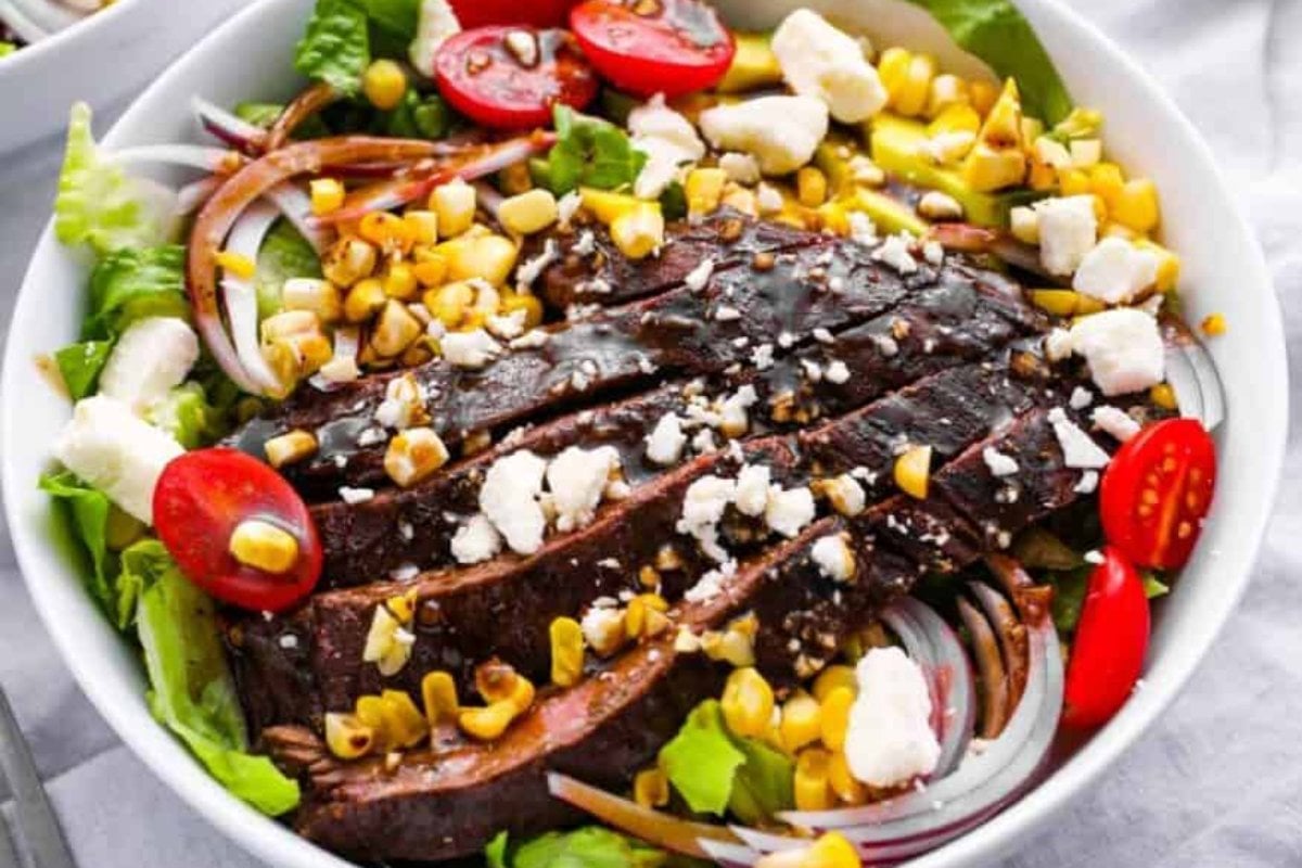A bowl of salad featuring grilled steak slices, cherry tomatoes, corn, red onions, lettuce, and feta cheese—a fresh addition to your favorite steak recipes.