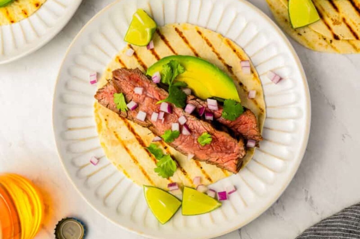 A grilled beef taco, inspired by popular steak recipes, is topped with chopped red onions, cilantro, and avocado slices and served on a white plate with lime wedges.