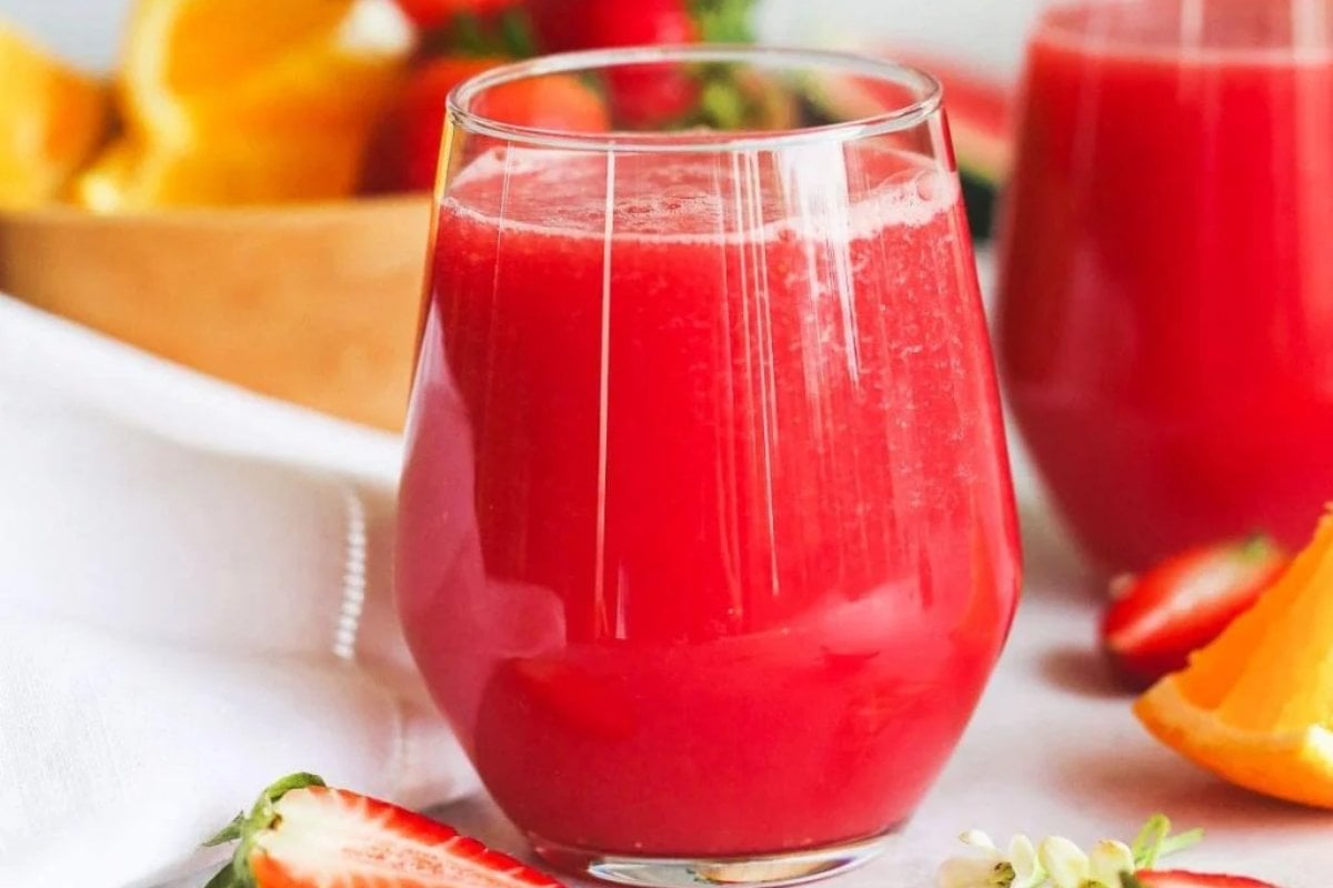 A glass filled with red fruit juice is placed on a table, surrounded by strawberry halves and orange slices. Another similar glass is in the background, reminiscent of refreshing watermelon recipes. The setting appears bright and fresh.