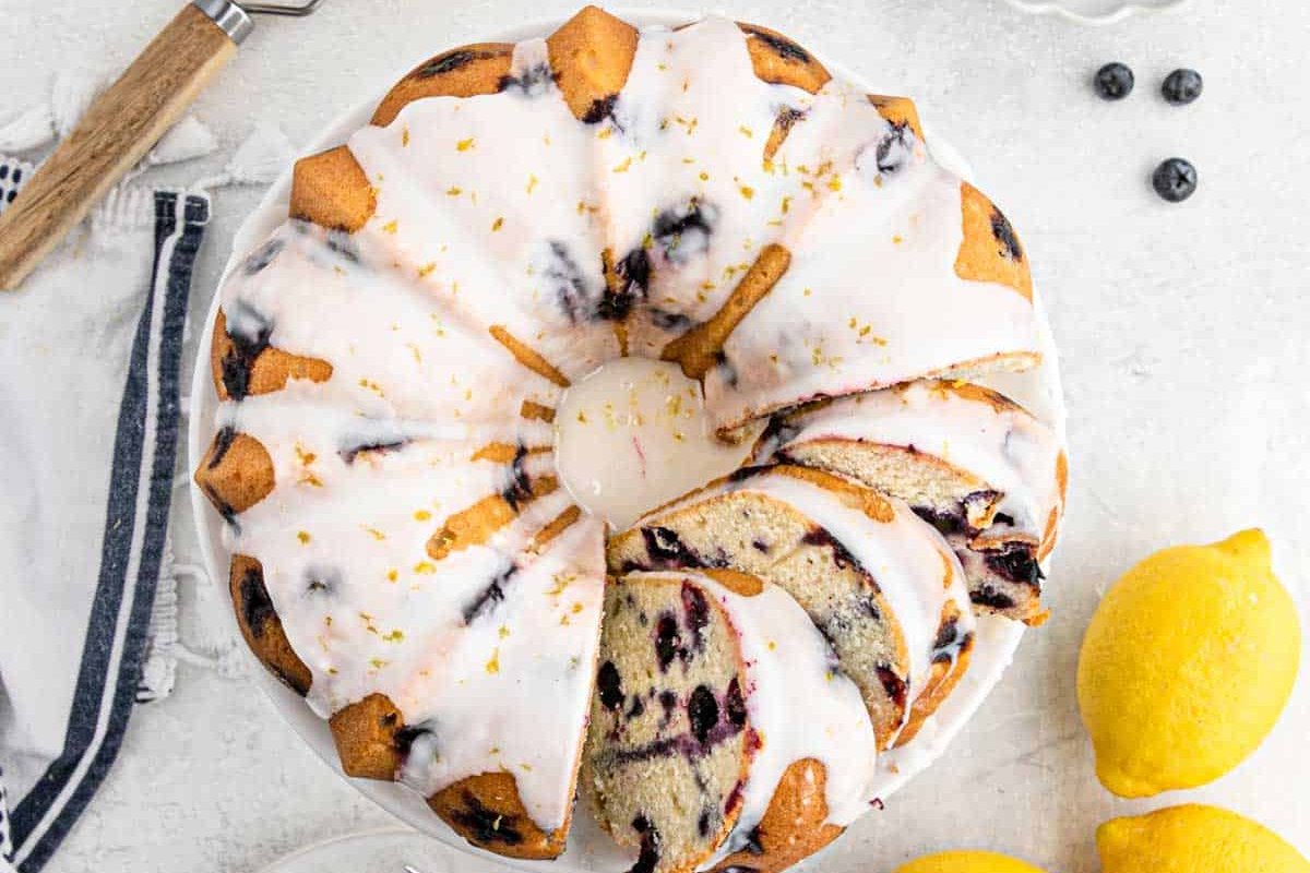 A delectable Bundt cake with white icing and blueberries, showcased on a white plate surrounded by lemons and a striped napkin&mdash;perfect inspiration for your new favorite blueberry recipes.
