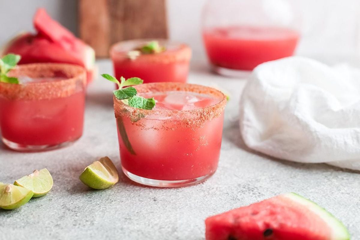 Four glasses of a red drink with ice and mint leaves sit on a table, rimmed with spice. Lime slices and watermelon pieces are scattered around, perfect for fresh summer watermelon recipes.