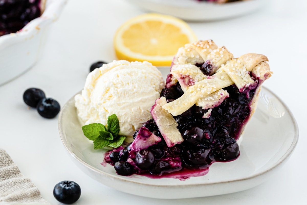 A slice of blueberry pie with a lattice crust is served on a white plate, showcasing one of the finest blueberry recipes, accompanied by a scoop of vanilla ice cream and garnished with a sprig of mint and a lemon slice in the background.