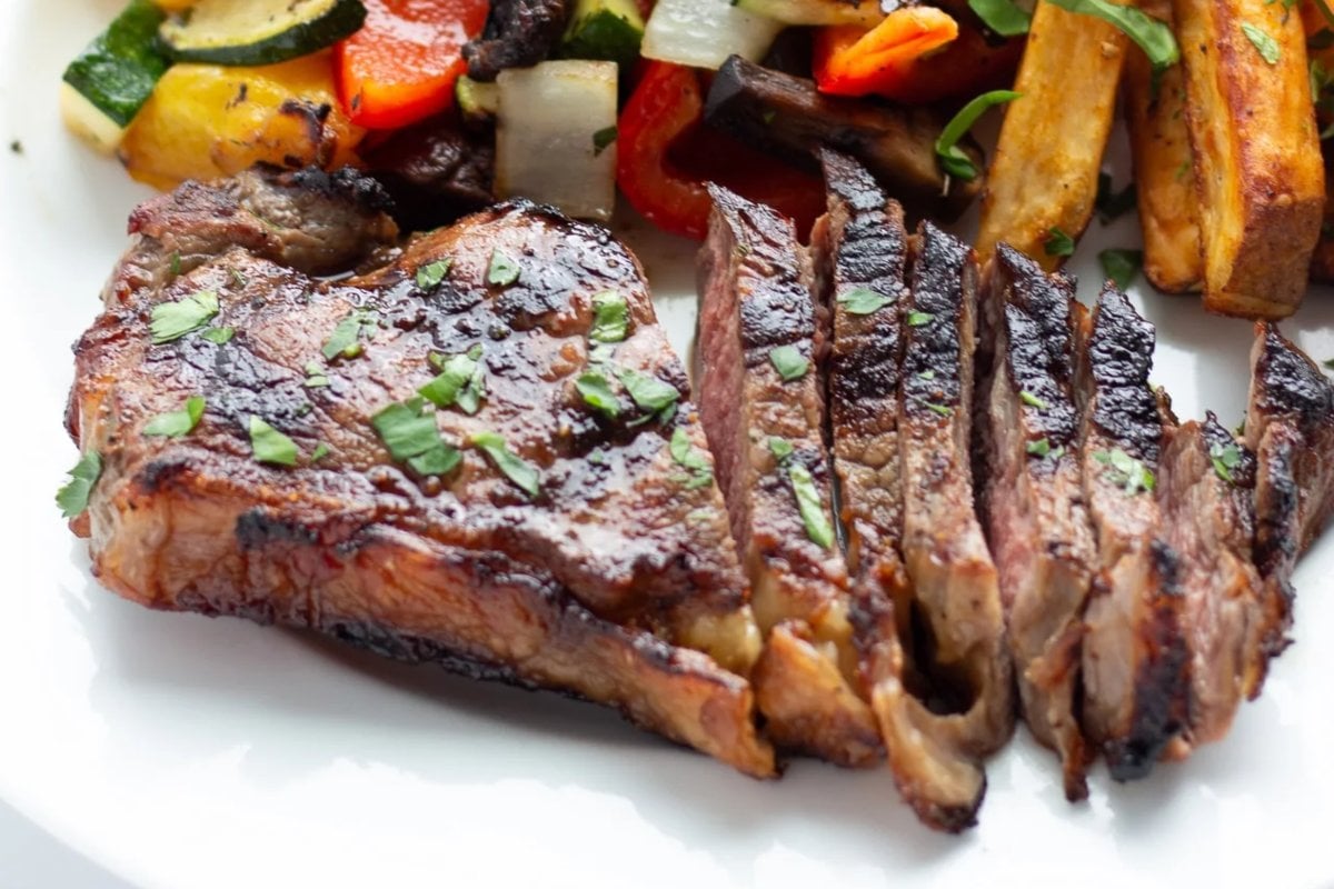 A grilled steak, sliced on a plate with roasted vegetables, garnished with chopped herbs, showcases one of the finest steak recipes.