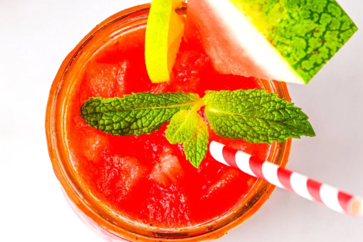 A close-up of a bright red beverage in a glass, garnished with a mint sprig, a slice of watermelon, and a slice of yellow lemon, with a red and white striped straw—a perfect addition to your favorite watermelon recipes.