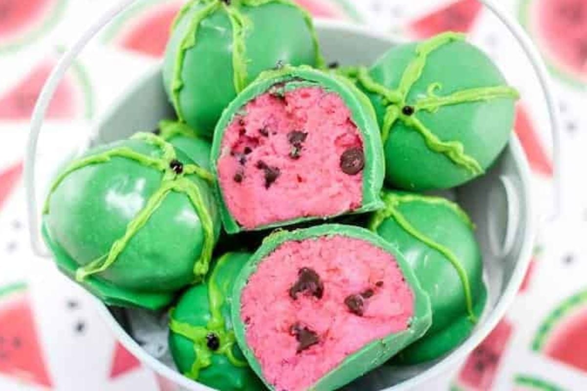 A bucket contains green watermelon-shaped cake pops with a pink interior and chocolate chip seeds, perfect for showing off your favorite watermelon recipes. Two cake pops are cut open to reveal their delightful insides.