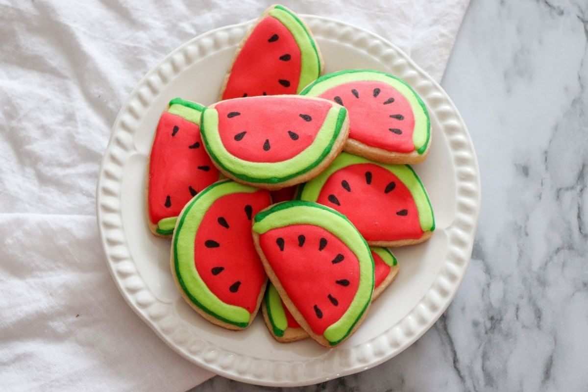 A white plate holds nine cookies decorated to look like watermelon slices, with red icing, green edges, and black seeds—perfect for those seeking creative watermelon recipes.