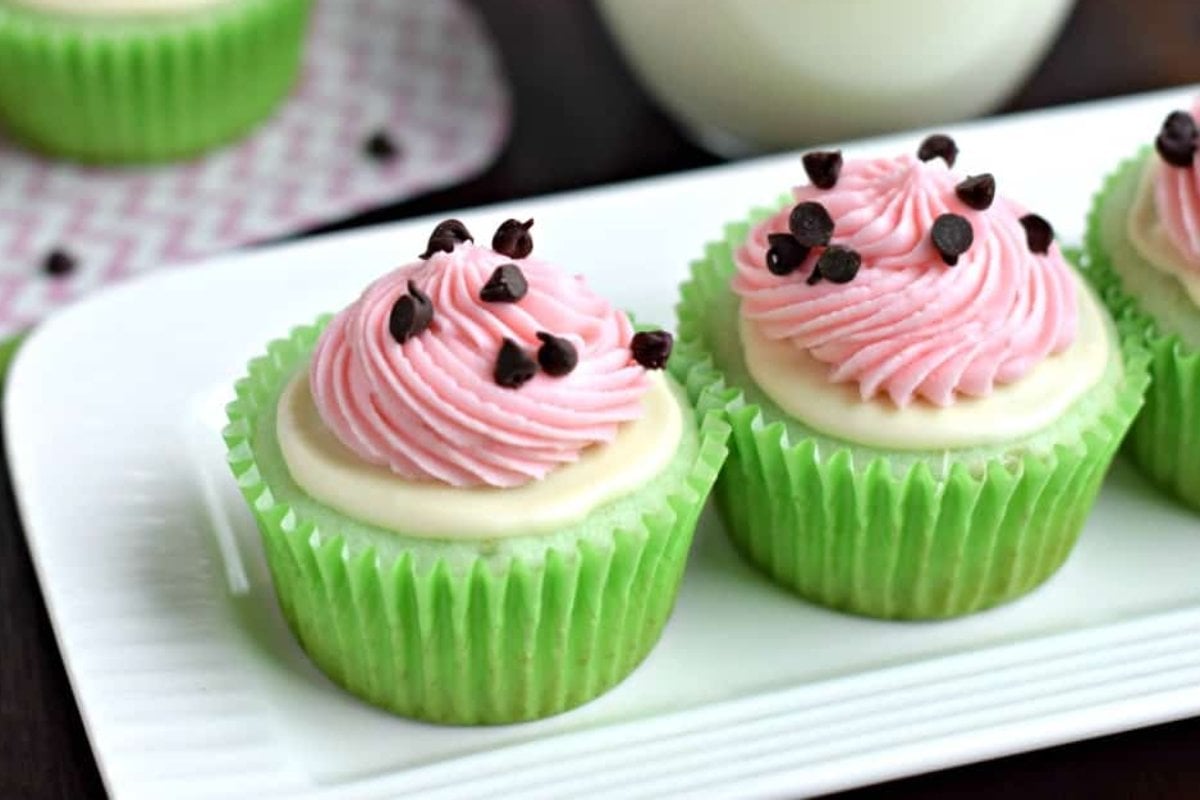 Three cupcakes with green wrappers, topped with pink frosting and small chocolate chips, reminiscent of delightful watermelon recipes, displayed on a white plate.