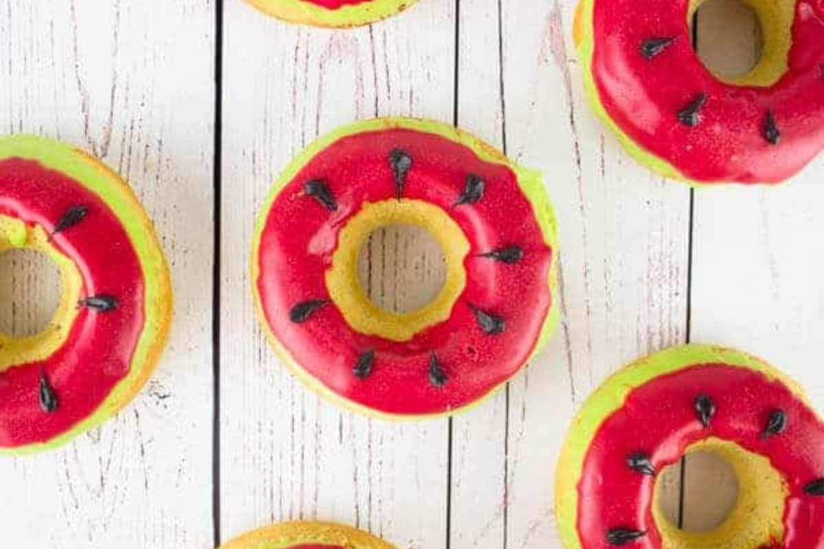 Donuts with red and green frosting resembling watermelon slices, topped with black seed-like decorations, displayed on a white wooden surface—perfect for adding a playful twist to your favorite watermelon recipes.