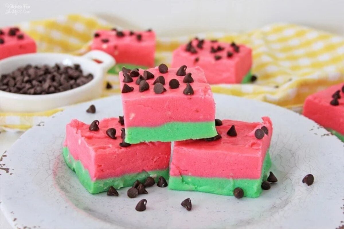 Three layered watermelon-themed fudge squares with pink and green colors, topped with chocolate chips, are arranged on a white plate. A bowl of chocolate chips and a yellow checkered cloth are in the background—perfect for adding to your collection of delicious watermelon recipes.