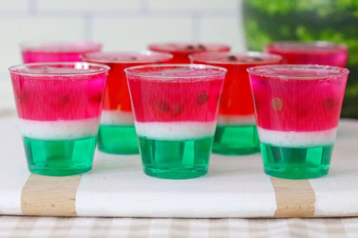 Watermelon-themed layered gelatin shots in clear plastic cups, featuring green, white, and pink layers with small chocolate chips as seeds, arranged on a white tray. These delightful treats make for perfect watermelon recipes to enjoy at any summer gathering.