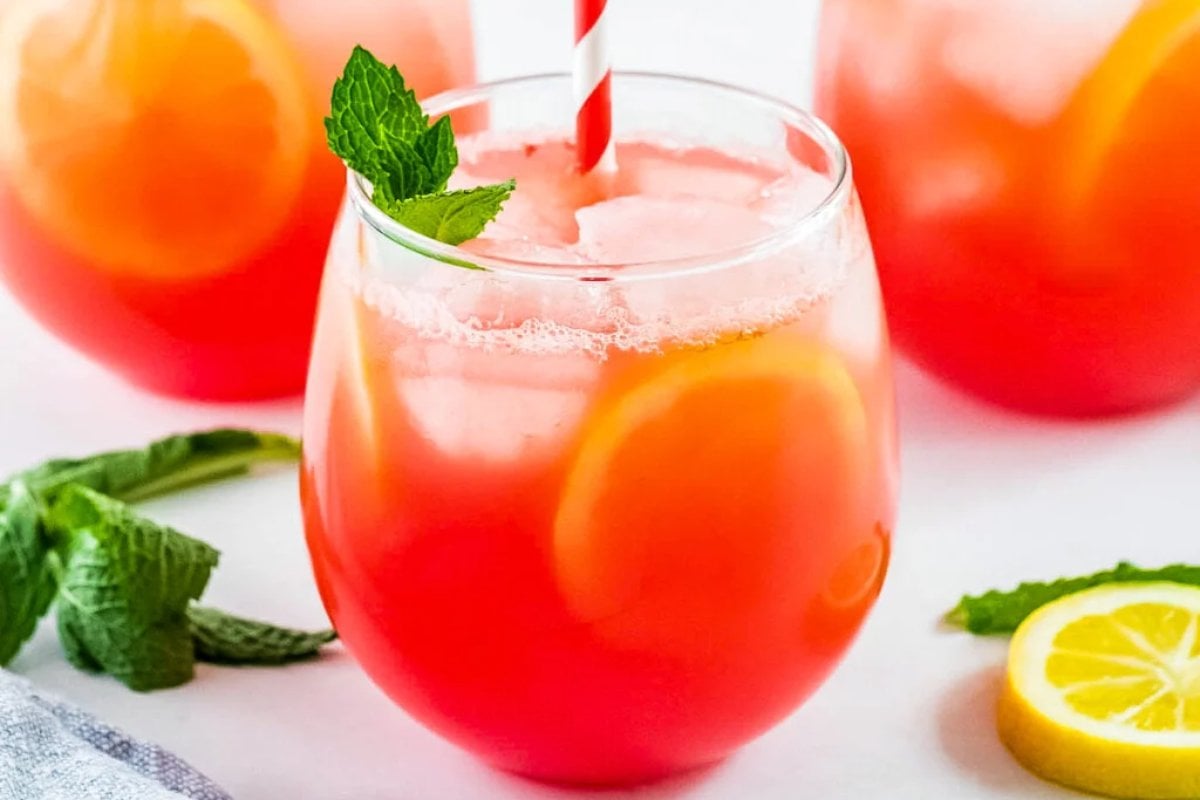 A refreshing glass of iced pink lemonade with a red and white striped straw, garnished with a mint sprig and a lemon slice, surrounded by additional lemonade glasses, lemon slices, and hints of watermelon recipes.