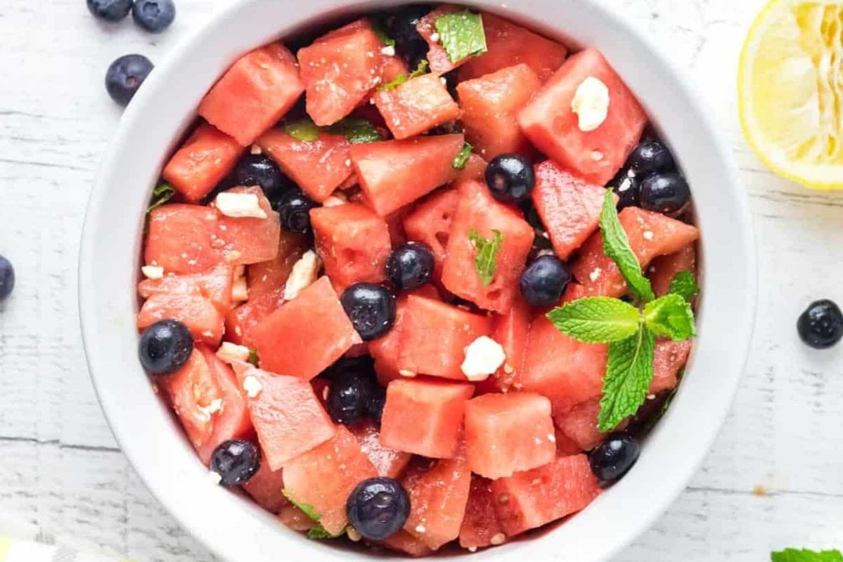 A bowl of watermelon cubes, blueberries, mint leaves, and small pieces of white cheese on a light wooden surface with a lemon wedge in the background—a delightful dish perfect for watermelon recipes.