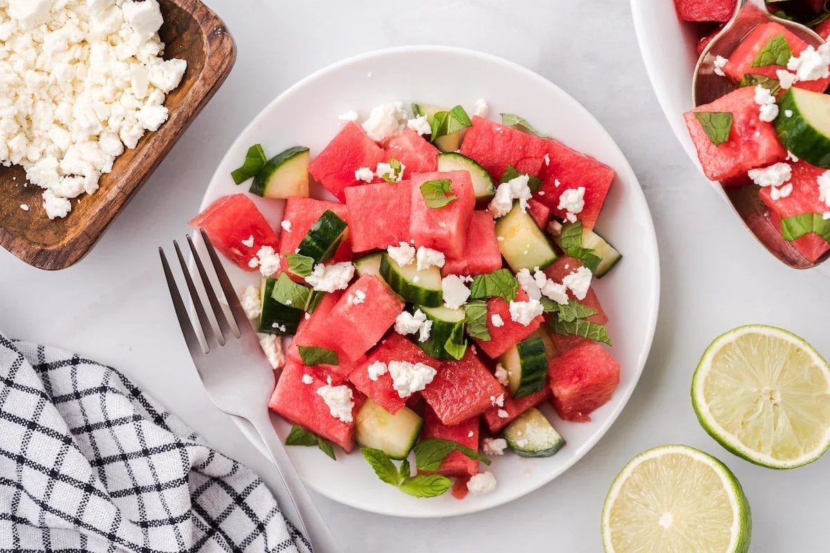 Plate of watermelon cucumber salad with feta cheese and basil on a white plate. Fork beside plate, sliced limes and a checkered napkin are also visible.