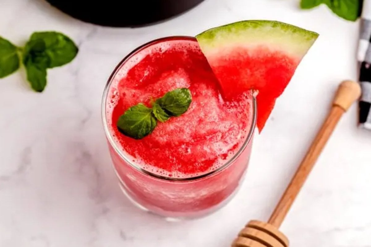 A glass of refreshing watermelon juice garnished with a slice of watermelon and mint leaves, placed on a marble surface next to a honey dipper and green leaves, perfect for your summer watermelon recipes.