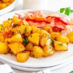 A white plate with seasoned, roasted air fryer breakfast potatoes garnished with parsley, accompanied by two strips of bacon. Nearby are parsley sprigs and a white and black checkered cloth.