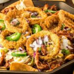 A skillet filled with BBQ chicken nachos topped with pulled pork, onion rings, sliced jalapeños, chopped red onions, and cheese. An embroidered cloth is partly visible on the right side.