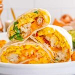 A plate with three breakfast burritos cut in half reveals a delicious mix of scrambled eggs, cheese, and hash browns. Surrounding the breakfast burritos are crispy tater tots and half an avocado.