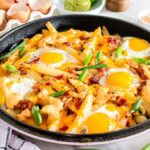 A skillet of breakfast fries topped with fried eggs, melted cheese, bacon, and green onions rests beside an empty egg carton, a dish of sauces, and a green lime on a kitchen countertop.