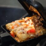 A pie iron on a grill contains a hot, melty dish of cheesy tater tots, diced tomatoes, onions, and melted cheese.
