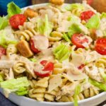 A bowl of chicken Caesar pasta salad with rotini, lettuce, cherry tomatoes, croutons, and shredded cheese, topped with creamy dressing.