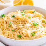 A bowl of creamy lemon pasta (also known as pasta al limone) garnished with grated cheese and parsley, with lemon halves and a salt shaker in the background.