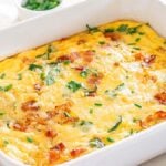A baked egg and hashbrown breakfast casserole in a rectangular white dish, garnished with chopped green herbs, and featuring visible pieces of bacon and spinach. This hash brown breakfast casserole is surrounded by several fresh ingredients, making it an inviting morning meal.