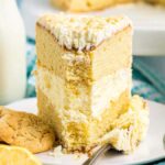 A slice of Italian lemon mascarpone cake with cream frosting on a plate, accompanied by cookies, lemon slices, and a small bowl of whipped cream. Two milk bottles are in the background.
