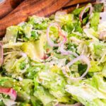 A bowl of fresh lemon parmesan chopped salad featuring lettuce, sliced red onions, and grated cheese, with wooden salad tongs resting on the side.
