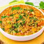 A bowl of lentil soup with vegetables, including carrots, tomatoes and celery, is placed on a wooden surface. Fresh ingredients surround the bowl of comforting lentil soup.