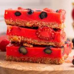A stack of mixed berry jello dessert bars with blueberries, strawberries and raspberries sits on a wooden board, with a bowl of strawberries and a striped cloth nearby.