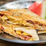 A metal plate with folded a pie iron breakfast crunchwrap filled with eggs, cheese, and bacon. A wedge of watermelon sits enticingly in the background.