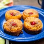 A blue plate with four individual pineapple upside-down cakes that were made in a pie iron on a campfire, each topped with a maraschino cherry, is set on a table beside a colorful cloth napkin, reminiscent of delightful treats made in a pie iron over an open flame.