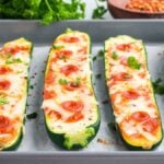 Four pizza zucchini boats topped with melted cheese, mini pepperoni slices, and chopped herbs are arranged on a baking sheet lined with parchment paper.