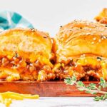 Close-up of juicy sloppy joe sliders with melted cheese and ground beef, topped with sesame seeds on buns, garnished with fresh thyme, and a checkered cloth in the background.