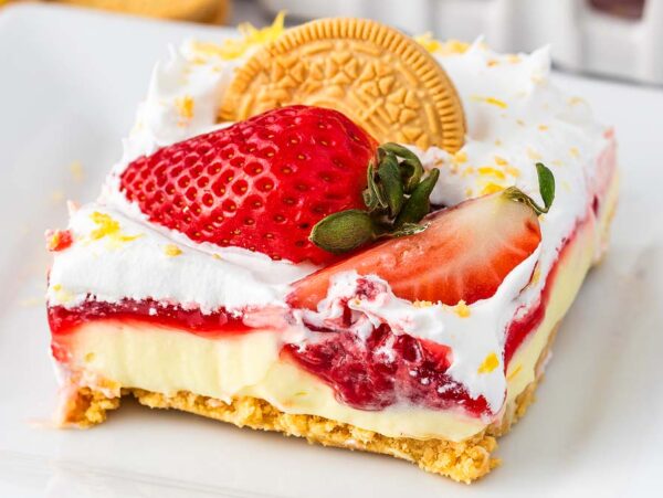 A square slice of indulgent strawberry lemon cheesecake lasagna featuring a cracker crust, a luscious cream filling, a tangy strawberry topping, whipped cream, and topped with a whole golden cookie and fresh strawberries.