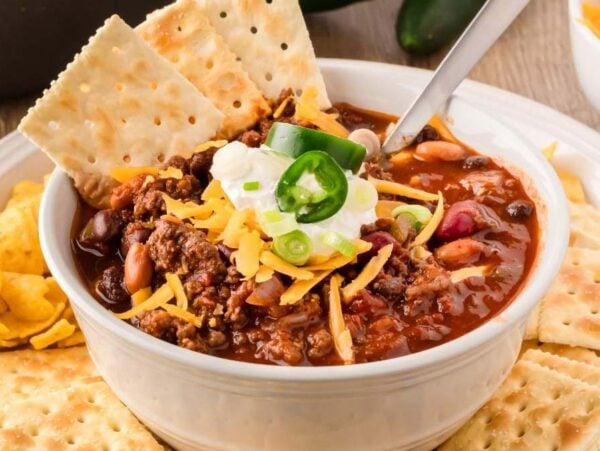 A bowl of venison chili topped with sour cream, shredded cheese, and sliced jalapenos, accompanied by saltine crackers and tortilla chips on a white plate.
