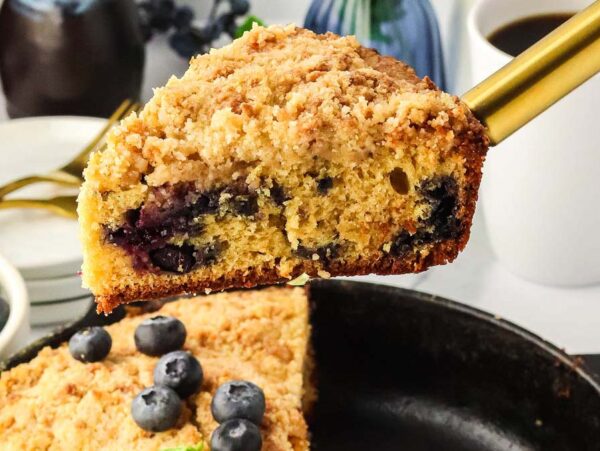 A slice of blueberry skillet crumb cake with visible blueberries is being served with a spatula. The cake is in a cast-iron skillet, and the background shows a cup of coffee and a bowl of blueberries.