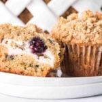A blueberry cream cheese muffin is sliced in half on a small white plate, revealing a luscious cream filling with a whole blueberry nestled inside. Another whole muffin sits invitingly next to it.