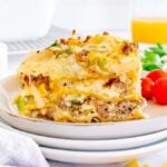 A piece of creamy, layered breakfast lasagna is served on a white plate, with cherry tomatoes and fresh greens in the background.