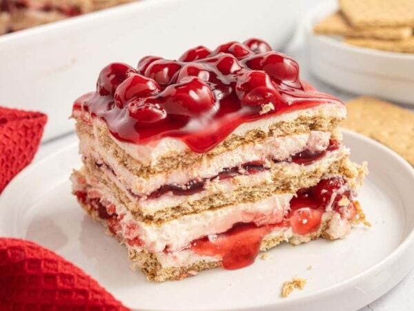 A slice of layered cherry cheesecake icebox cake on a white plate, featuring graham crackers, creamy filling, and cherry topping. A red cloth and graham crackers are in the background.
