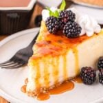 A slice of creme brûlée cheesecake topped with blackberries, and whipped cream, served on a white plate with a fork. A mint leaf and additional blackberries are on the side.