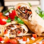 A close-up of a sliced Halifax donair wrap on a wooden board. The wrap contains seasoned meat, diced tomatoes, onions, and other fillings. Fresh tomatoes and parsley garnish the scene.