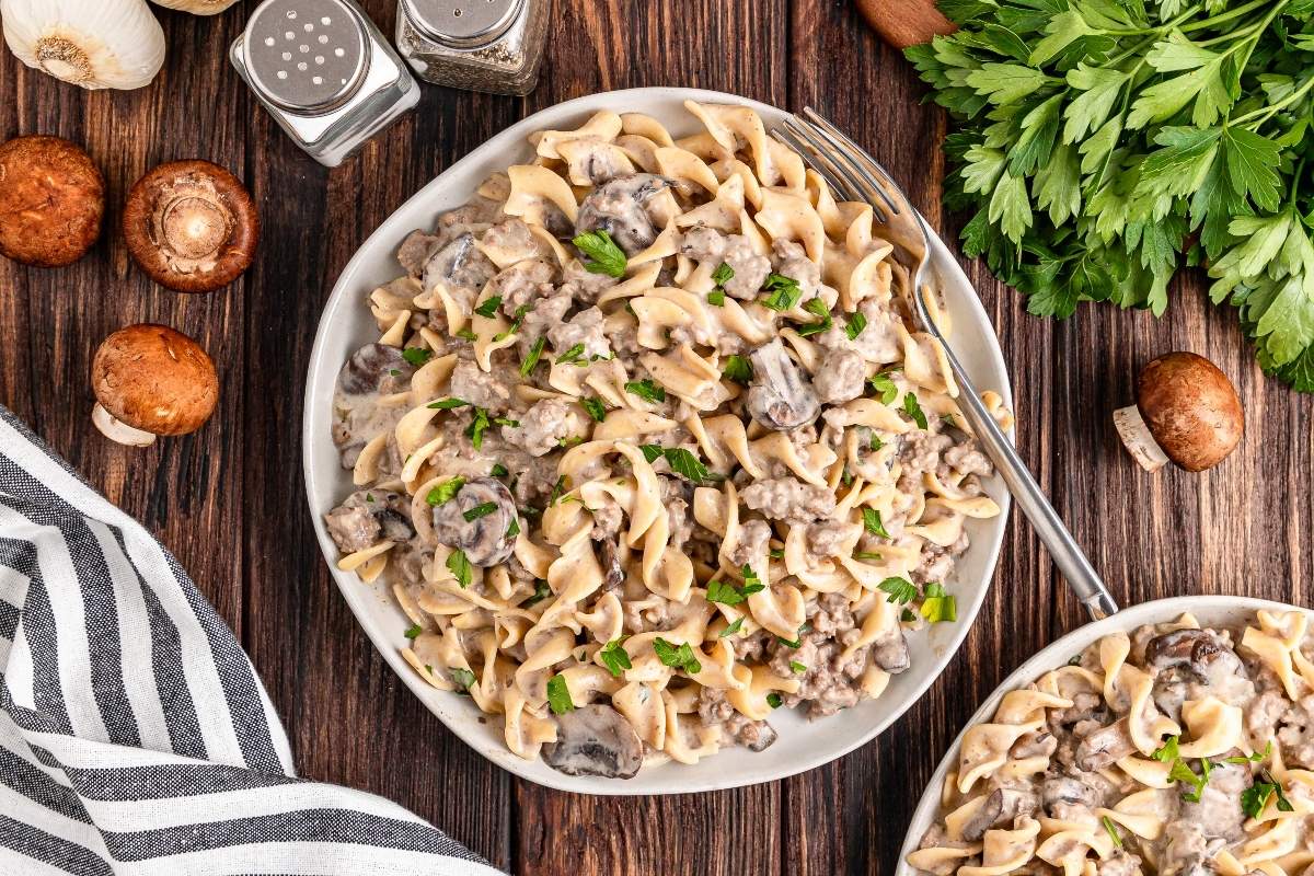 A plate of pasta with creamy mushroom sauce and chopped parsley, surrounded by mushrooms, parsley, salt and pepper shakers, and a striped cloth on a wooden table—reminiscent of a refined ground beef stroganoff.