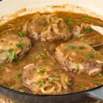 Four Hamburger Steaks with Homemade Gravy and onions in brown gravy, garnished with parsley, in a large skillet with a spoon.