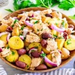 A bowl of Italian tuna potato salad topped with chunks of chicken, olives, red onion slices, and fresh parsley sits on a table with a cut lemon and a bowl of extra olives nearby.