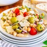 A plate of Italian white bean salad with cherry tomatoes, olives, shaved cheese, and herbs sits surrounded by a bowl of cherry tomatoes, slices of bread, and a glass container of olive oil.