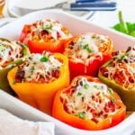 A white baking dish filled with colorful Italian stuffed peppers topped with melted cheese, garnished with chopped herbs. A fork, knife, and pepper grinder are in the background.