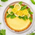 Lemon tart with a golden crust, garnished with lemon slices and fresh mint leaves, displayed on a white plate and surrounded by whole lemons and mint leaves.