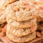 A stack of homemade oatmeal butterscotch cookies (also known as oatmeal scotchies) is placed on a wooden board, with several more cookies and butterscotch chips scattered in the background.