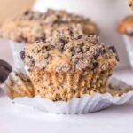A close-up of Oreo muffins with crumbled chocolate cookies on top, placed on a table. Some muffins have their liners partially peeled back, and a stack of chocolate sandwich cookies is in the background.